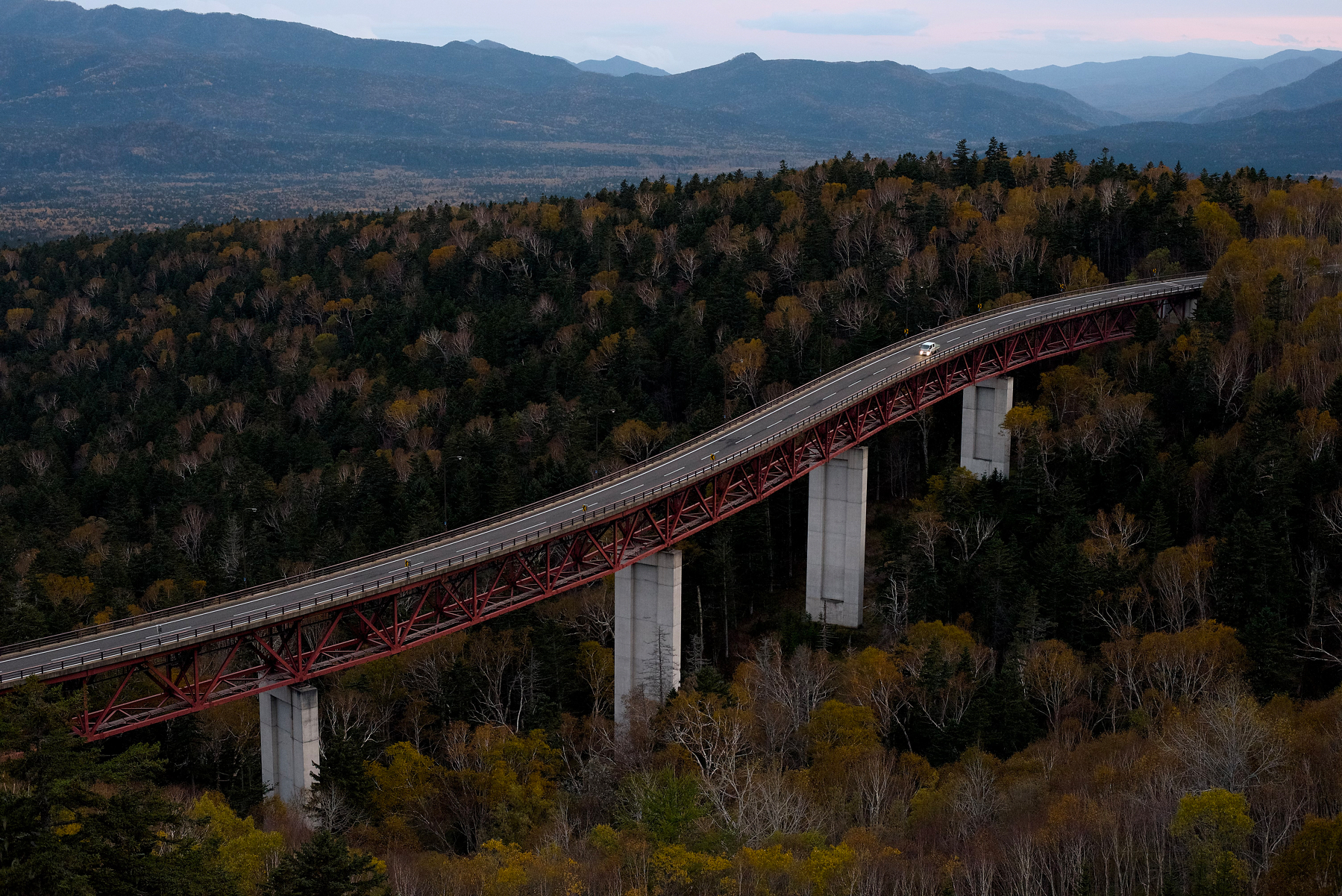 A long, elevated bridge stretches over a dense forest with mountains in the background.